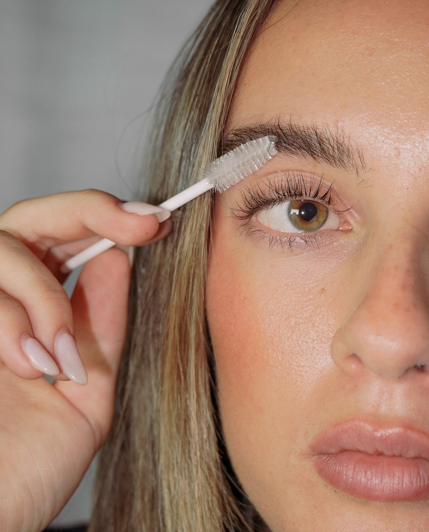 A close-up of a woman holding a spoolie eyebrow makeup brush against her eyebrow, showing her face and hand in detail after an at-home lash lift. 