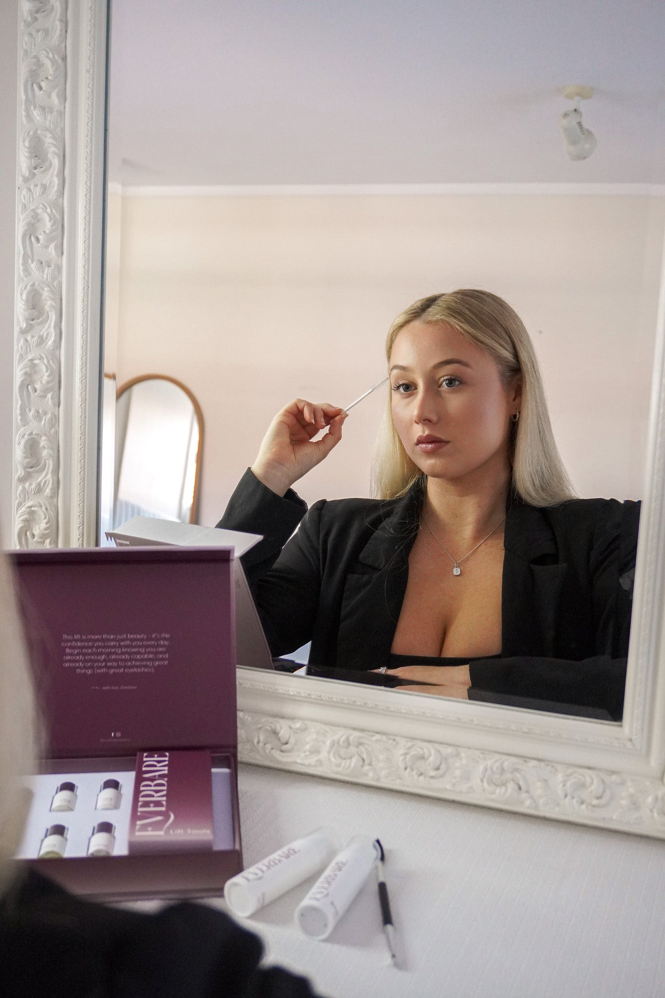 A woman with blonde hair looks into a large ornate white mirror while applying mascara wand for an lash lift. In the foreground, an open purple cosmetic box labeled 'EVERBARE' and two white cosmetic tubes rest on a white vanity table.